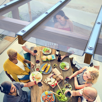 People sitting around a wooden table with food, viewed through a glass roof.