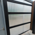 Metal gate with glass panels and a dark frame, part of a larger fence system.
