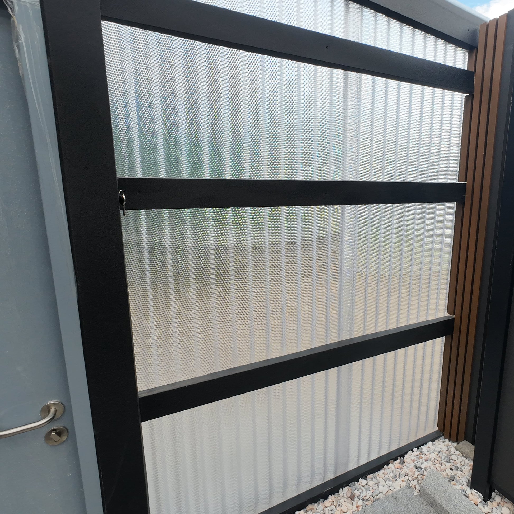 Metal gate with glass panels and a dark frame, part of a larger fence system.